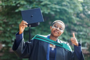 Positive young black lady with short black hair in academic regalia standing and holding diploma giving thumbs up
