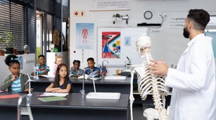Students in a classroom listen to their teacher, who holds a model skeleton.