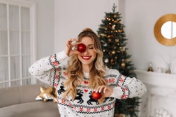 Portrait of a young cheerful woman with red lips and curly hair in a knitted sweater laughing and holding a red Christmas ball covering her eyes with it against the background of a Christmas decorated Christmas tree at a holiday in December at home