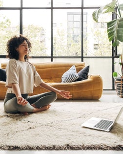 Young multiracial latina woman meditating at home with online video meditation lesson using laptop.