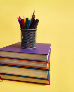 A metal pencil holder with school supplies on stacks of multicolored books. Teacher's Day concept, literary, knowledge, Back to School, education. Yellow background copy space