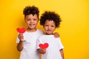 Portrait of two cute positive small brothers cuddle hold red paper heart empty space isolated on yellow color background