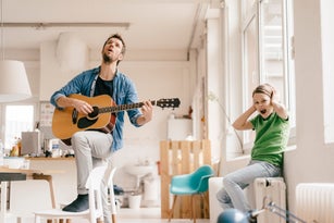 Horrified son covering his ears with father playing guitar at home