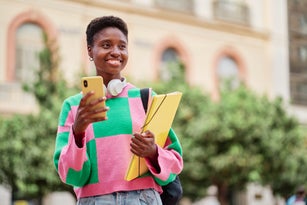Waist-up view of young cheerful African American student woman smiling with backpack and folder while using her mobile phone in the city street.