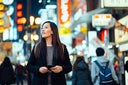 Young Asian female traveller with backpack searching for directions with smartphone GPS navigation app while exploring and strolling in busy downtown city street at night in Osaka, Japan. Lifestyle and technology. Travel, vacation and holiday concept