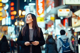 Young Asian female traveller with backpack searching for directions with smartphone GPS navigation app while exploring and strolling in busy downtown city street at night in Osaka, Japan. Lifestyle and technology. Travel, vacation and holiday concept