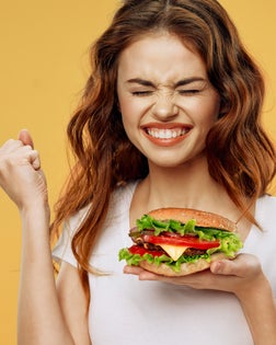 A young woman in a white T-shirt holds a big burger in one hand and shows the Yes gesture with the other hand.