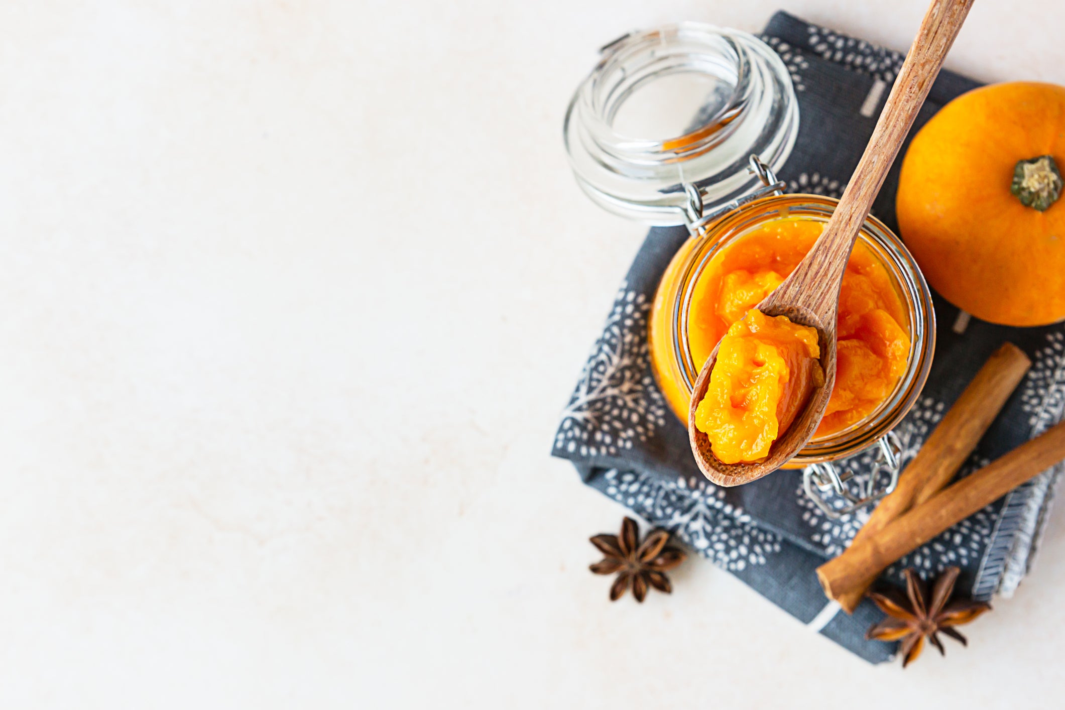 Organic pumpkin puree in glass jar and wooden spoon with fresh pumpkin and spices on linen napkin. Light concrete background. Top view.