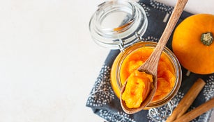 Organic pumpkin puree in glass jar and wooden spoon with fresh pumpkin and spices on linen napkin. Light concrete background. Top view.