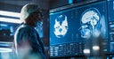 Medical Hospital Research Laboratory: Caucasian Male Neurosurgeon Looking At TV Screen With Brain MRI Scans Of Patient, Analyzing, Preparing For Complicated Neurosurgery, Wearing Surgical Gown.