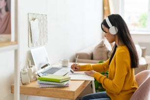 Adult learning online at home. Handwriting in notebook while listening to lecture in headphones. Asian female improving knowledge at remote college course. Concept of gaining new skills from internet. Covid-19 pandemic distancing, side view