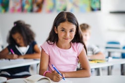 A small school girl sitting at the desk in classroom, looking at camera.