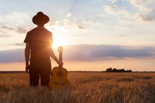 Musician holding acoustic guitar and walking in summer fields, sunset