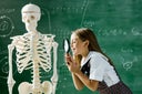 girl in a classroom standing in front of a chalkboard examining a human skeleton with a magnifying glass