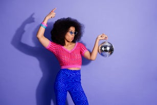 Cheerful fashionable woman in retro outfit posing with disco ball on vibrant violet background, embracing vintage trends