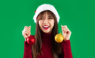 Young pretty asian woman in red sweater wearing santa hat posing smiling and holding red and gold glitter balls on green screen background.