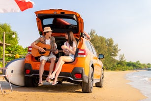 Beautiful young couple sitting behind the car with acoustic guitar at beach near sea