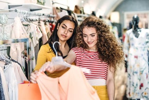 Two female teenager friends standing inside in the shop, holding and looking at dress.