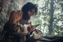 Primeval Caveman Wearing Animal Skin Holds Sharp Stone and Makes First Primitive Tool for Hunting Animal Prey, or to Handle Hides. Neanderthal Using Handax. Dawn of Human Civilization