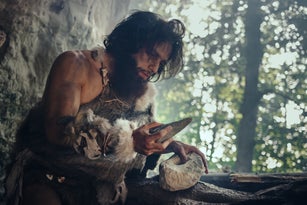 Primeval Caveman Wearing Animal Skin Holds Sharp Stone and Makes First Primitive Tool for Hunting Animal Prey, or to Handle Hides. Neanderthal Using Handax. Dawn of Human Civilization