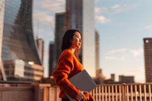 Confident young Asian businesswoman with laptop walking in the city against skyscraper