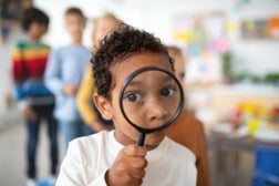 Pre- school tboy playing with magnifying glass indoors at nursery school, looking at camera.