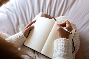 POV high angle view of Unrecognizable young woman wearing a robe sitting on bed writing on journal in cozy bedroom.