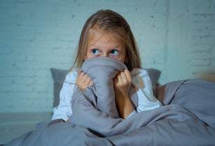 Scared little girl staying sleepless hiding behind the duvet looking horrified in the dark having childhood nightmares in child imagination Sleeping disorders Stress Depression and Insomnia concept.
