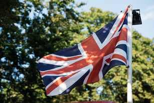 Union Jack flag of United Kingdom proudly waves on a bright sunny day, casting shadows on its fabric. Green trees provide a natural, leafy background