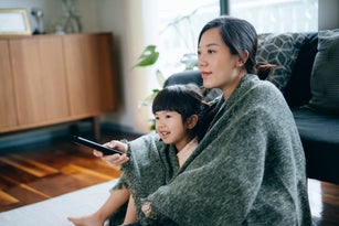 Loving young Asian mother and cute little daughter wrapped in blanket, relaxing at home and watching TV in the living room. Bonding between mother and daughter. Family lifestyle. Love and care concept