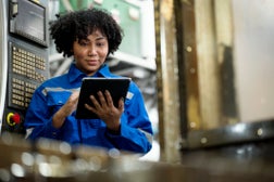 CNC milling machine setup process. Female African American Mechanical Engineer prepares the machine and engineering workpiece base on the machining standard. She is using a tablet in a production line.