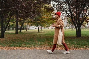 Mid adult woman with mobile phone and reusable shopping bag walking outside in the city on autumn day.