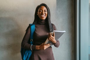 Portrait of cheerful young woman holding book standing with backpack against gray wall