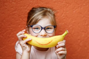 Happy little child girl with yellow banana like smile on orange background. Preschool girl with glasses smiling. Healthy fruits for children