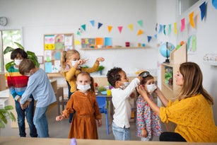 Pre school teacher helping little girl to wear face mask indoors at nursery school, coronavirus concept.