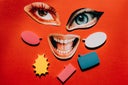 A flat lay conceptual collage featuring cutouts of expressive human eyes and a wide-open smiling mouth on a vibrant red background.