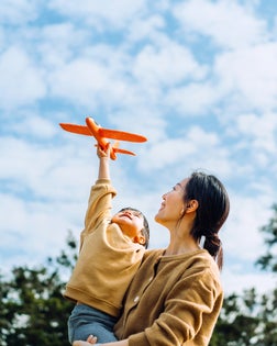 Young Asian mother and lovely little daughter spending time together outdoors, playing with airplane toy and smiling joyfully in park on a lovely sunny day against beautiful blue sky