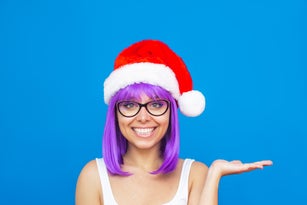 Young woman in glasses with violet hair and red Santa hat holding hand presenting empty copy space