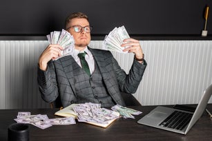 A young serious man in a gray formal suit and glasses sits at his desk and counts cash. A businessman holds money.
