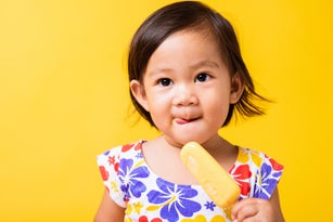 Happy portrait Asian baby or kid cute little girl attractive laugh smile wearing dick pattern shirt holds and eating sweet wooden ice cream, studio shot isolated on yellow background, summer concept