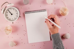 Female hand writing plans, goals in white note pad on pink background. Directly above