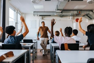 Rear view of male and female junior high students with hand raised while teacher teaching in classroom at school