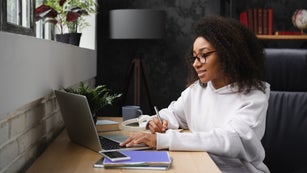 African young teenager e-learning on distance, using laptop, writing homework assignment in copybook, watching webinars online from home.