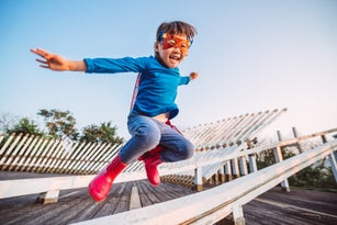 Cheerful little girl with a handmade superhero disguise jumping over wooden fence joyfully in park