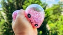 Pink glitter antistress bao toy with kawaii face held outdoors against green foliage background, sparkling squishy stress relief