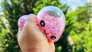 Pink glitter antistress bao toy with kawaii face held outdoors against green foliage background, sparkling squishy stress relief