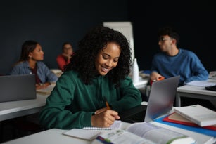 A female student smiling as she writes on a notebook in a classroom.