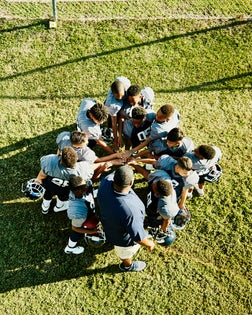 Overhead view of coach and football team gathered in circle with hands together before football game