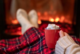 Woman with cup of hot cocoa and marshmallow warming legs in winter white socks near fireplace flame, covered christmas plaid
