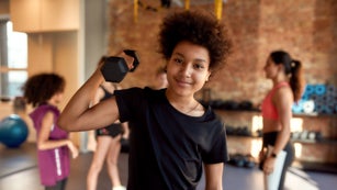 Kid smiling at camera while exercising using dumbbell in gym.